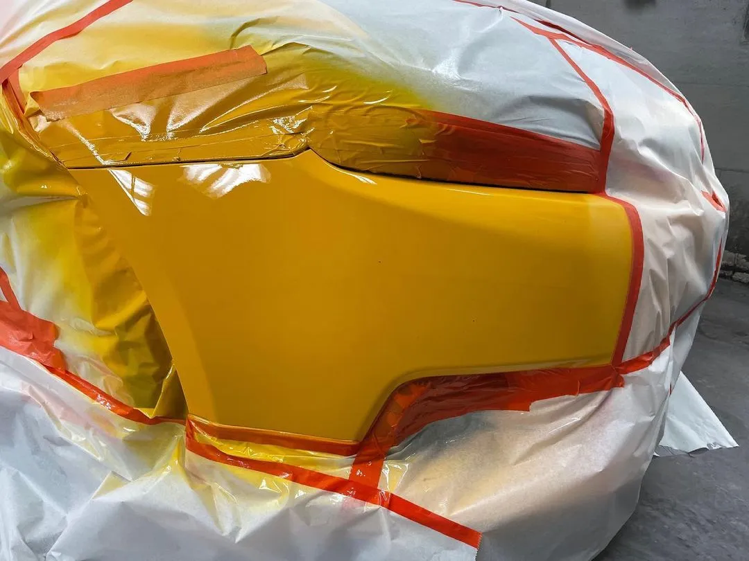 A section of a car freshly painted in yellow, with careful application of protective plastic on areas to prevent unintended paint, showcasing meticulous attention to detail in the painting process.