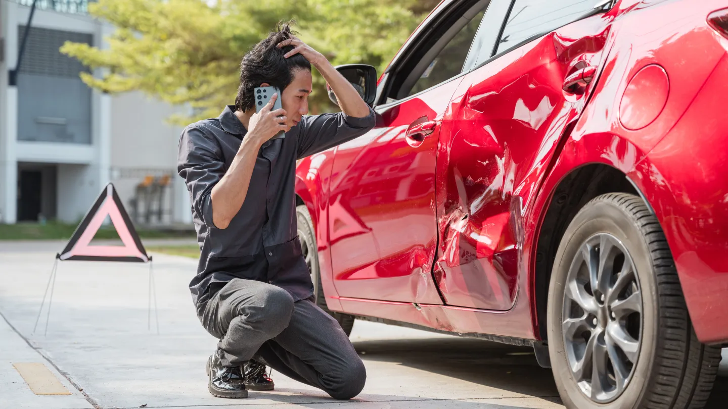 Driver kneeling next to a damaged red car calling for help after a collision