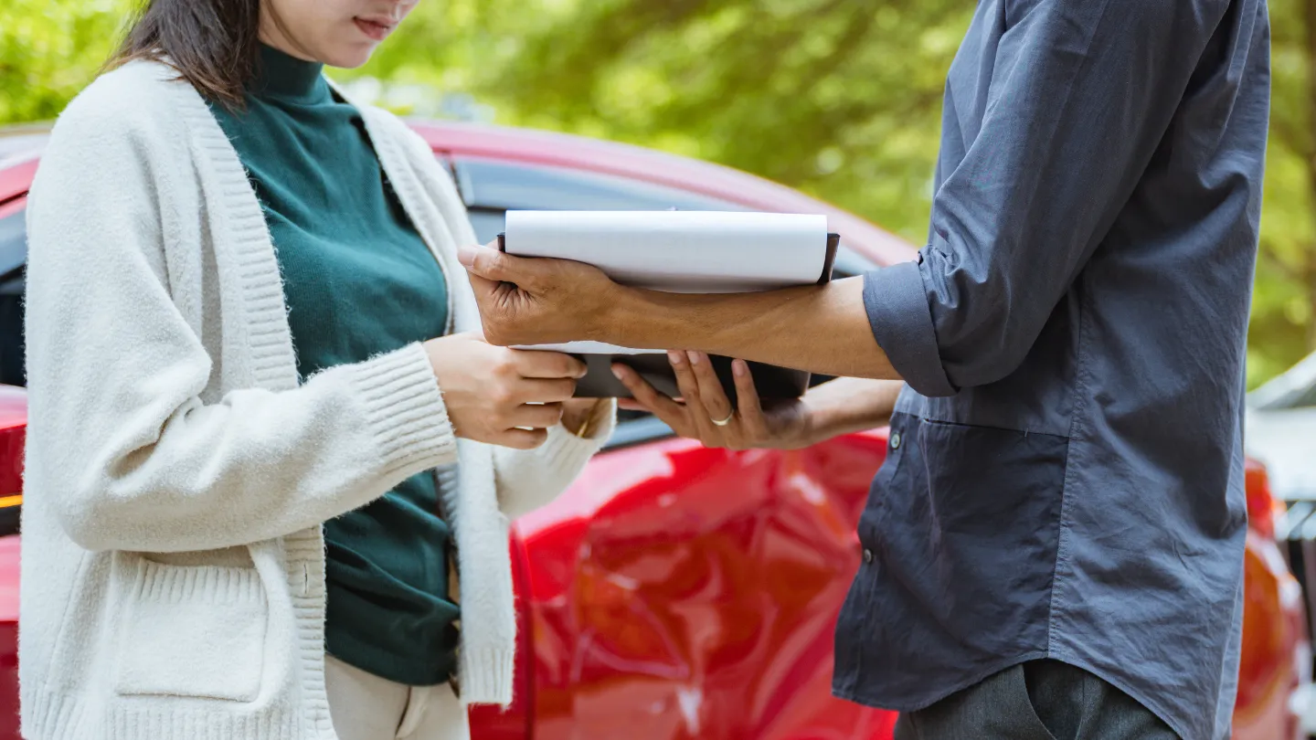 Two people exchanging insurance documents in front of a damaged red car