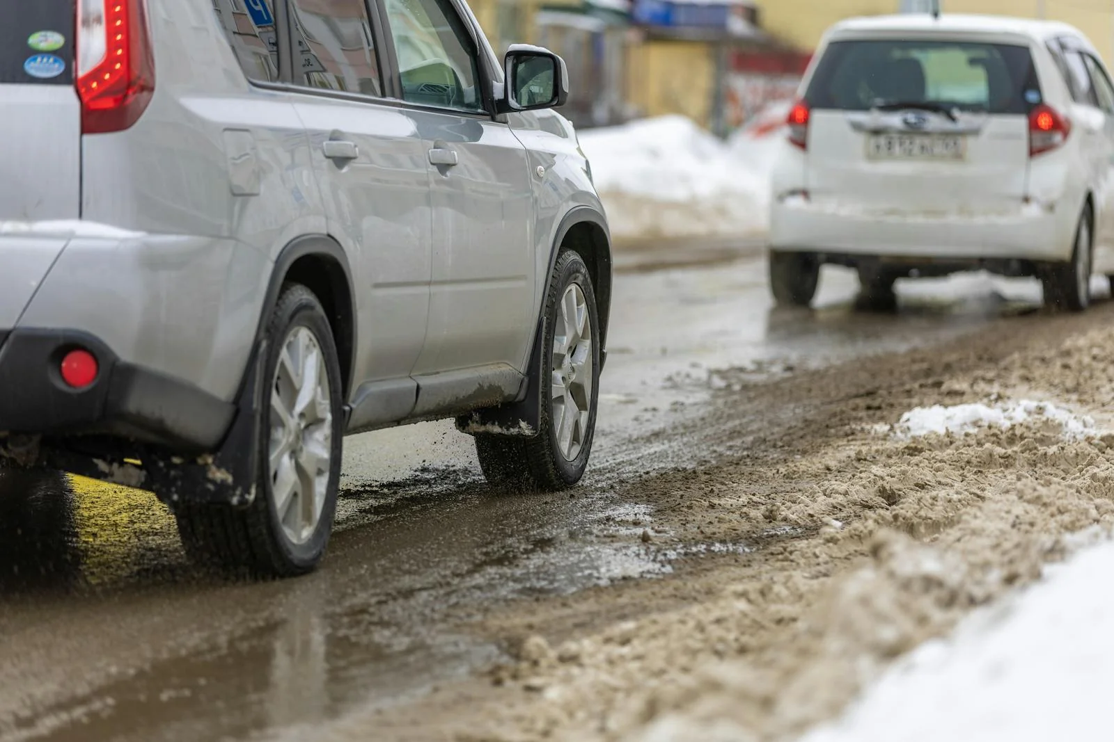 Cars driving on a slushy winter street with salt residue and snow piled along the curb