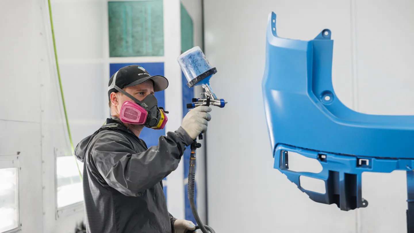 Best Auto Body technician spray-painting a blue bumper cover in a paint booth